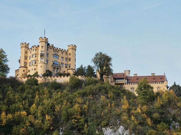 Vor einem klaren blauen Himmel liegt auf einem mit grünen und gelben Bäumen bewachsenen Hügel das Schloss Hohenschwangau mit mehreren Türmen.